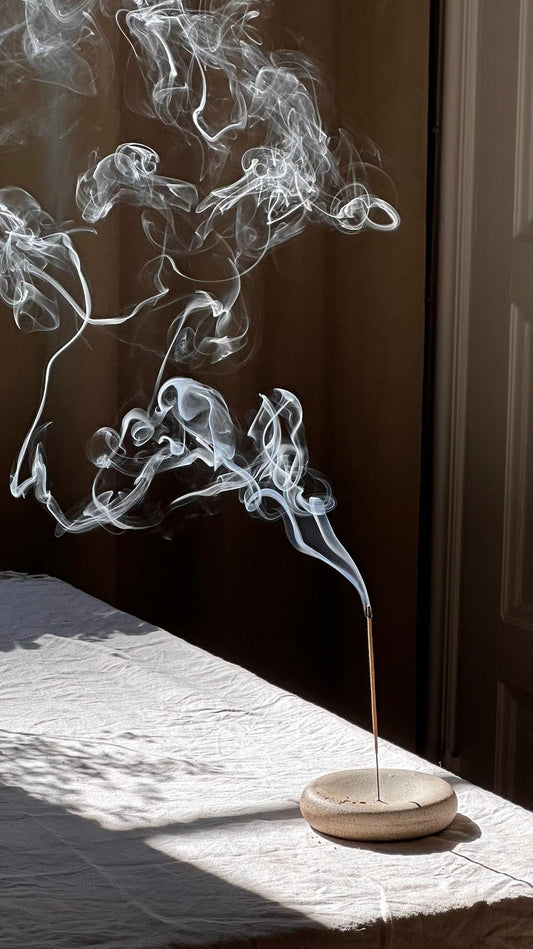 A hand lighting a stick of incense in a small stone holder on a bright windowsill