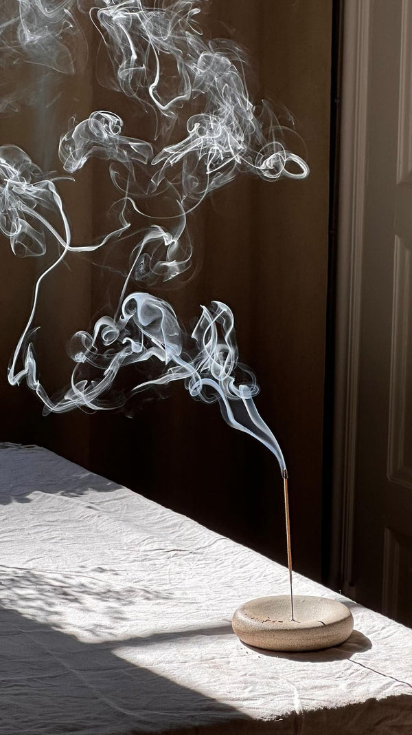 A hand lighting a stick of incense in a small stone holder on a bright windowsill