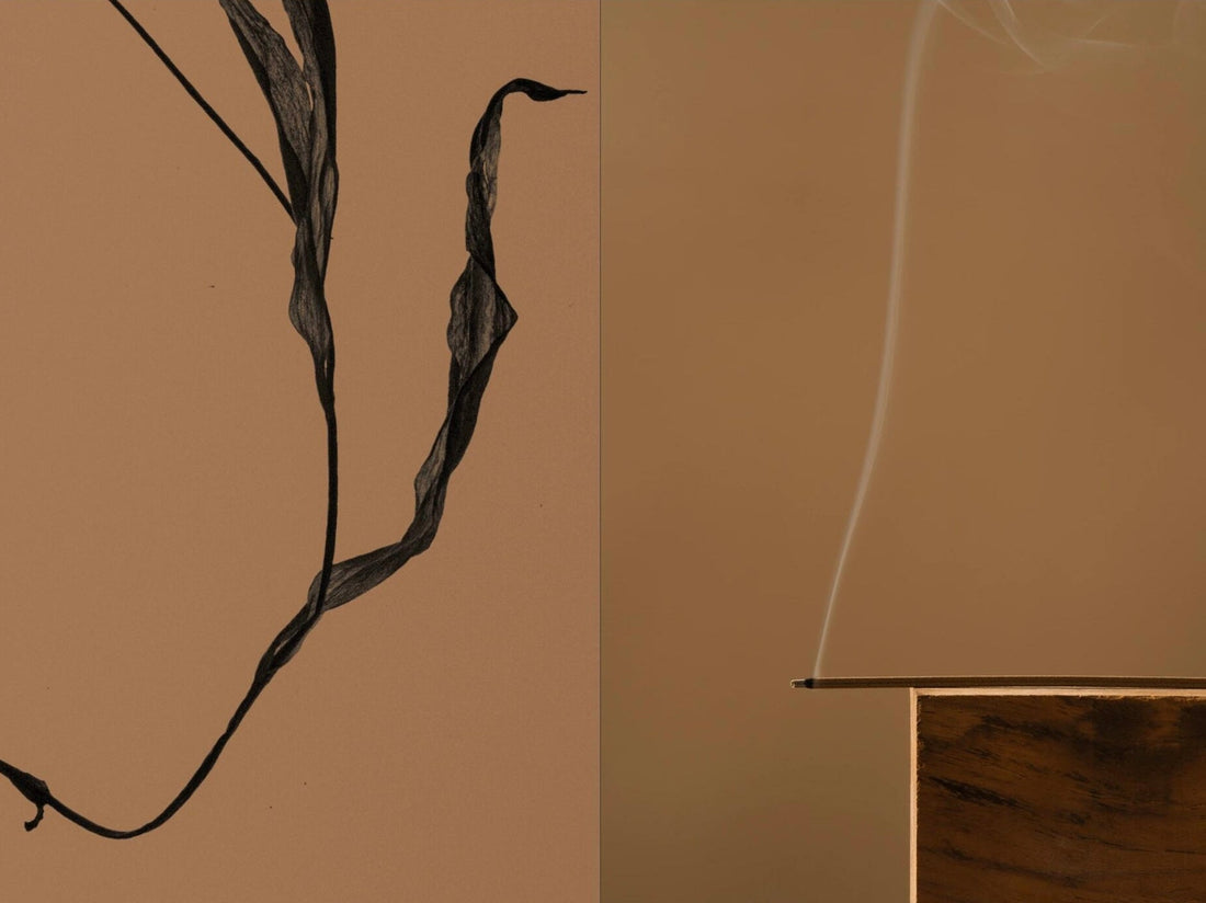 A dried curling leaf beside a lit incense stick balanced on a wooden block, a thin ribbon of smoke rising from its tip