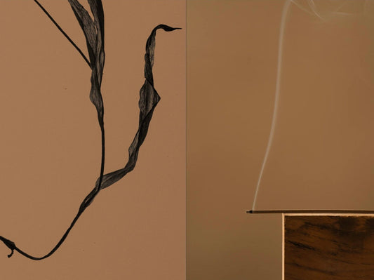 A dried curling leaf beside a lit incense stick balanced on a wooden block, a thin ribbon of smoke rising from its tip