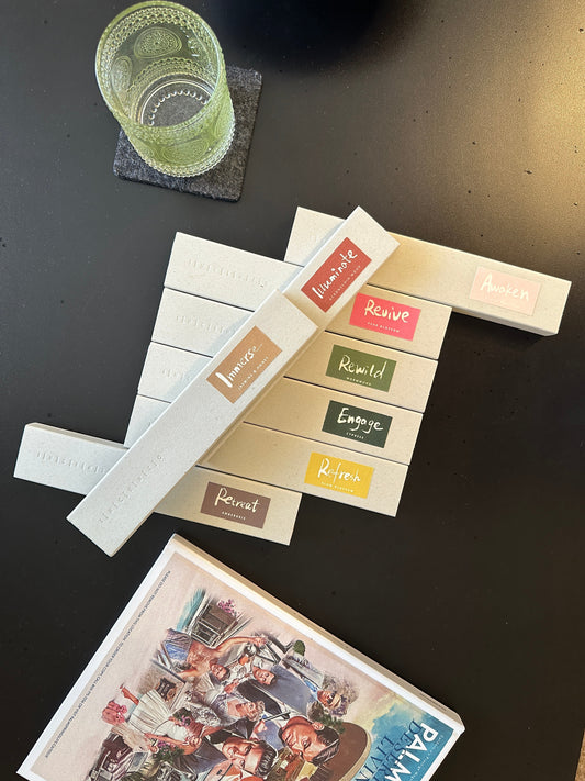 INCENZO incense boxes fanned out on a dark table beside a green glass tumbler, labels reading Illuminate, Revive, Rewild, Engage, Refresh, Retreat and Immerse